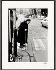 An elderly woman with a cane stands at the edge of a crosswalk on a cobblestone street. She wears a dark coat and scarf, cars and buildings in the background. This black and white scene makes an evocative fine art print for conscious interior design.