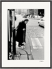 An elderly woman with a cane stands at the edge of a crosswalk on a cobblestone street. She wears a dark coat and scarf, cars and buildings in the background. This black and white scene makes an evocative fine art print for conscious interior design.