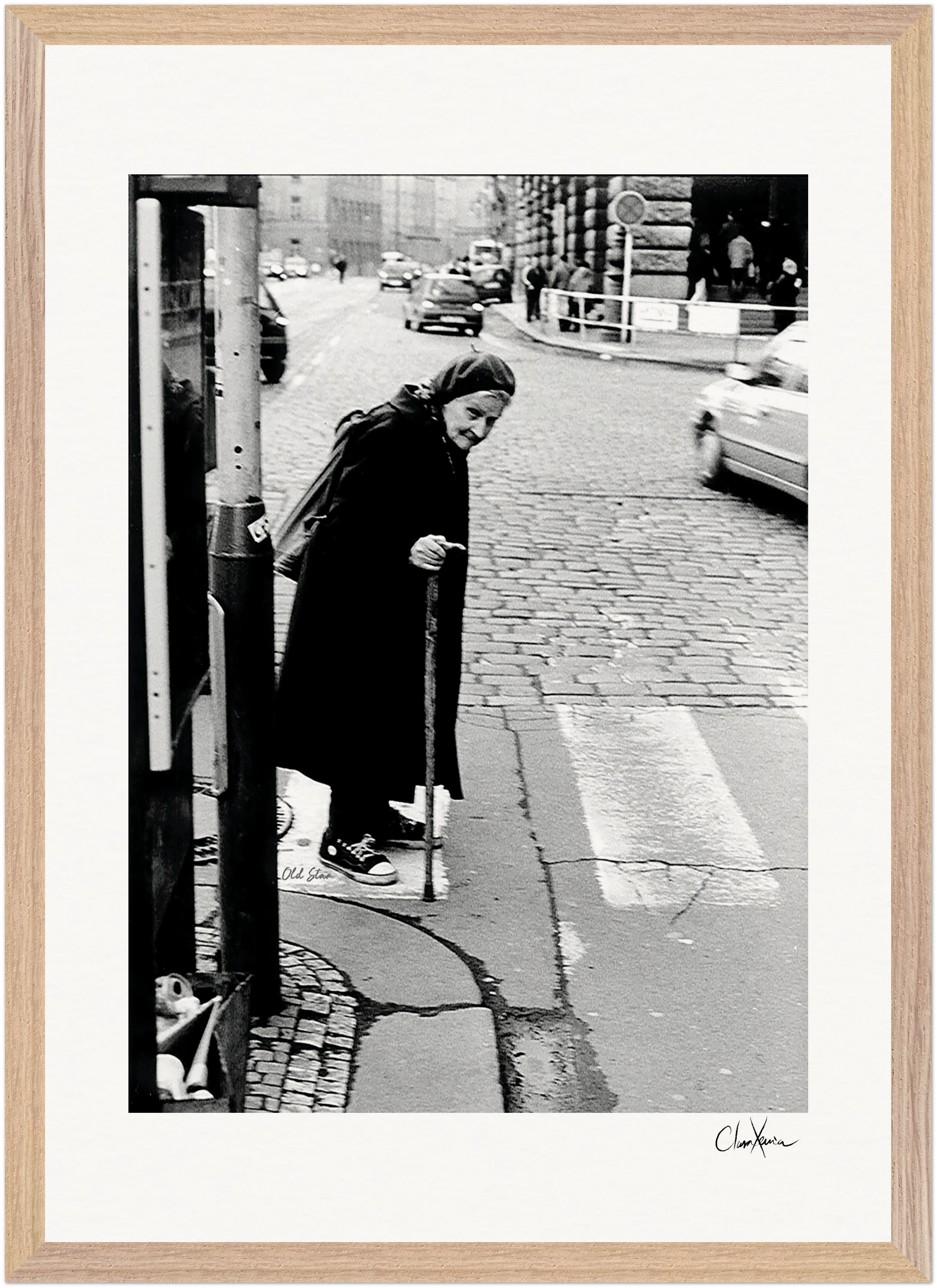 An elderly woman with a cane crosses a cobblestone street at a crosswalk, moving slowly in dark clothing. Cars and buildings line the background. This black and white photo adds an inspirational wall art touch to conscious interior design.