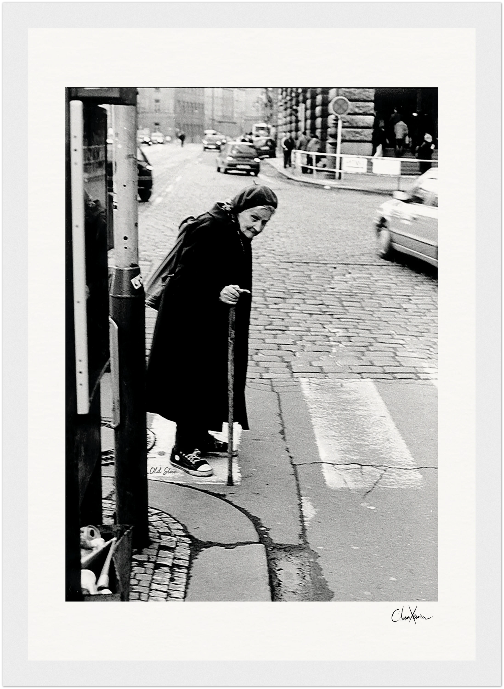 An elderly woman with a cane stands at a crosswalk on a cobblestone street, wearing a long coat and headscarf. Cars appear in the background. The black-and-white scene makes elegant framed wall art for conscious interior design.