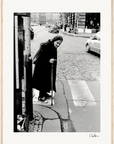 An elderly woman in a dark coat and headscarf leans on a cane while crossing a cobblestone street. Cars and pedestrians linger in the background. This black-and-white scene is perfect as inspirational wall art or a fine art print.