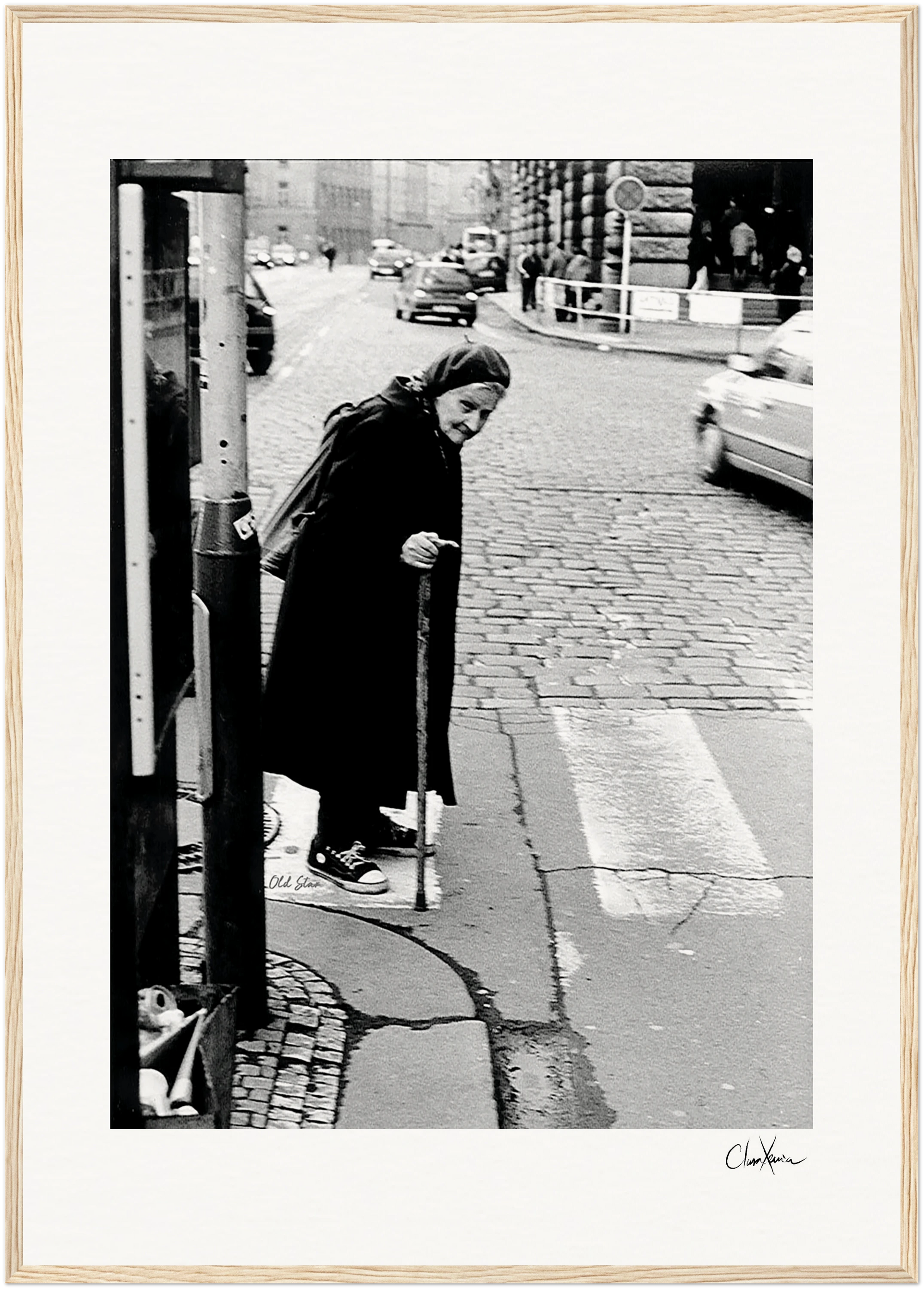 An elderly woman in a dark coat and headscarf leans on a cane while crossing a cobblestone street. Cars and pedestrians linger in the background. This black-and-white scene is perfect as inspirational wall art or a fine art print.