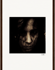 A dark, grainy portrait of a person’s face, partially obscured by shadows and strands of hair, is centered on a white background and framed in brown wood—an excellent choice for mindful home decor. A signature appears in the lower right corner.