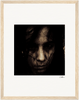 A close-up, grainy portrait of a person with dark hair partially covering their face, set against a black background and framed in a gallery-quality frame. Their expression appears serious and intense—perfect for mindful home decor or as a fine art print.