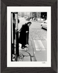 An elderly woman with a cane stands at a crosswalk on a cobblestone street as cars pass by. This black-and-white photograph, set in a gallery-quality frame, is perfect as mindful home decor or striking framed wall art.