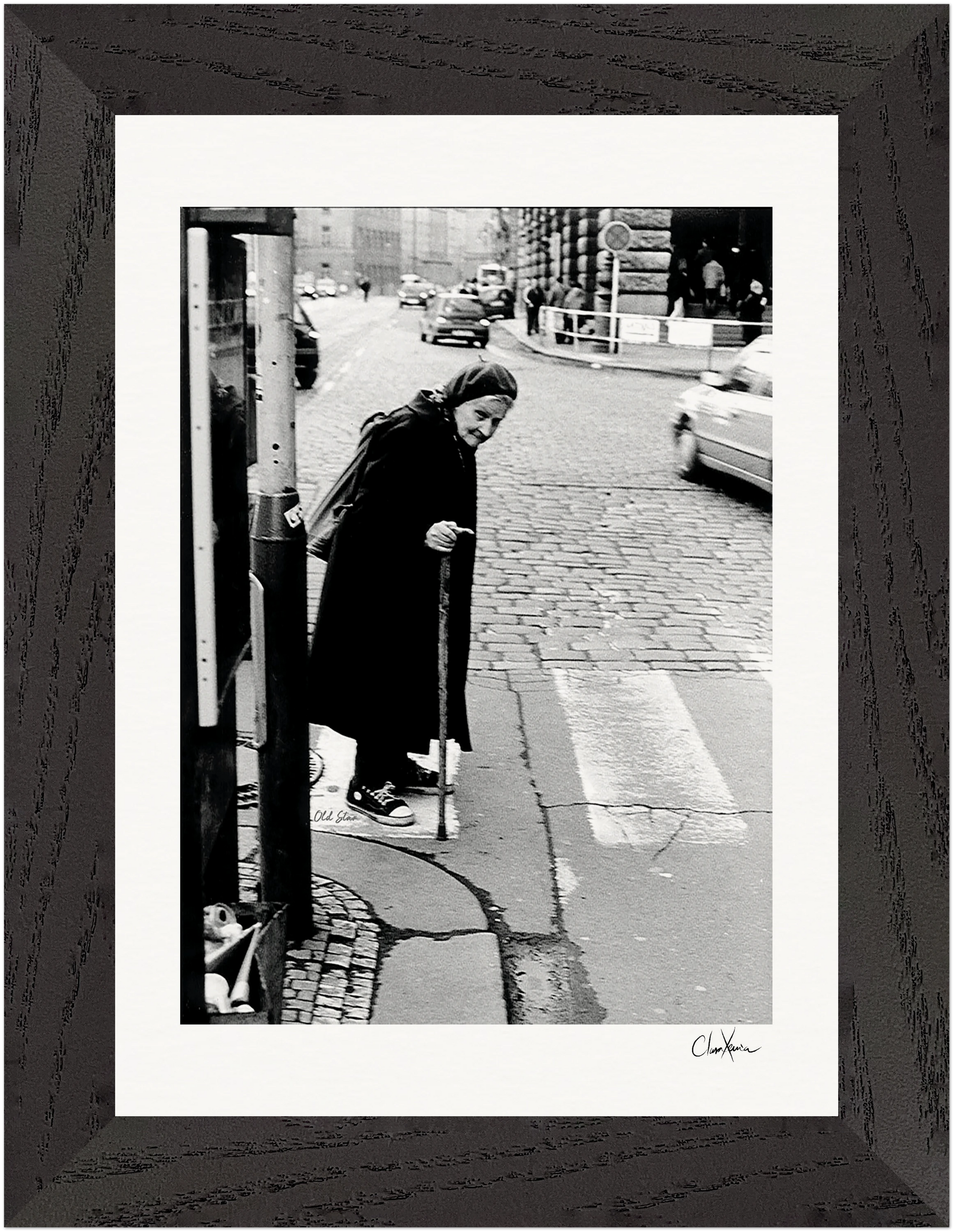 An elderly woman with a cane stands at a crosswalk on a cobblestone street as cars pass by. This black-and-white photograph, set in a gallery-quality frame, is perfect as mindful home decor or striking framed wall art.