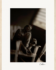 A man sits on a wooden chair indoors, one knee drawn up, gazing thoughtfully into the distance. This sepia-toned inspirational wall art features soft lighting, a blurred background, and a signature at the bottom right.