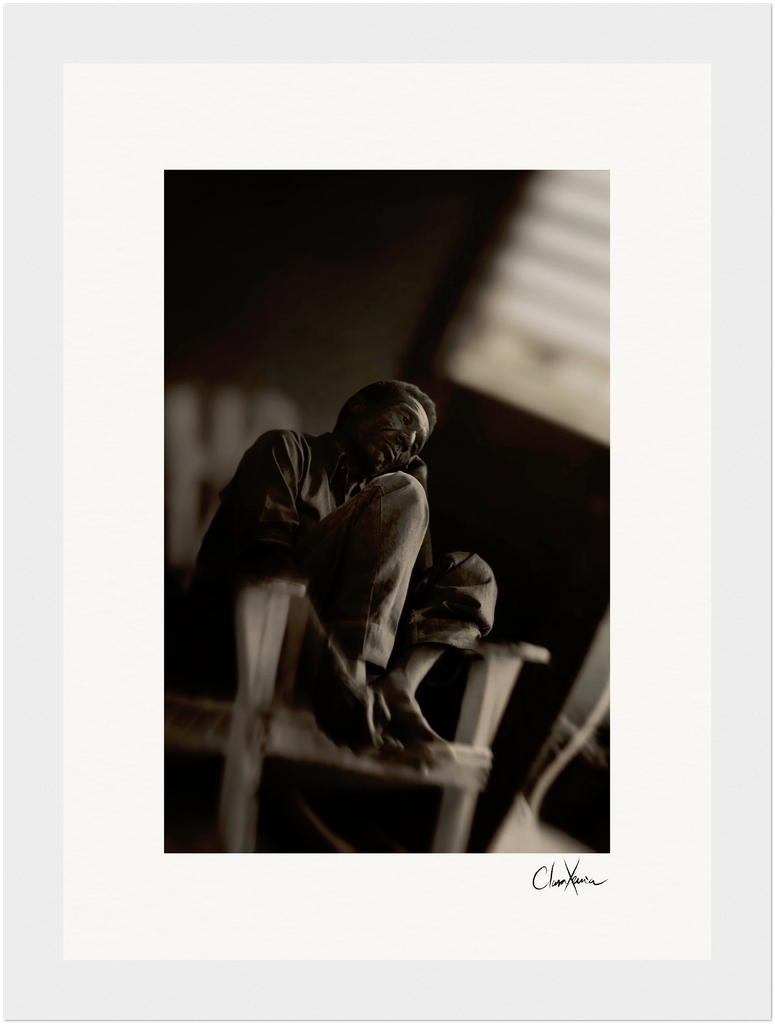 A black-and-white photo of a person sitting on a chair indoors, resting their chin on their hand with one leg pulled up. The blurred, dimly lit background enhances the conscious interior design, making it perfect as inspirational wall art.