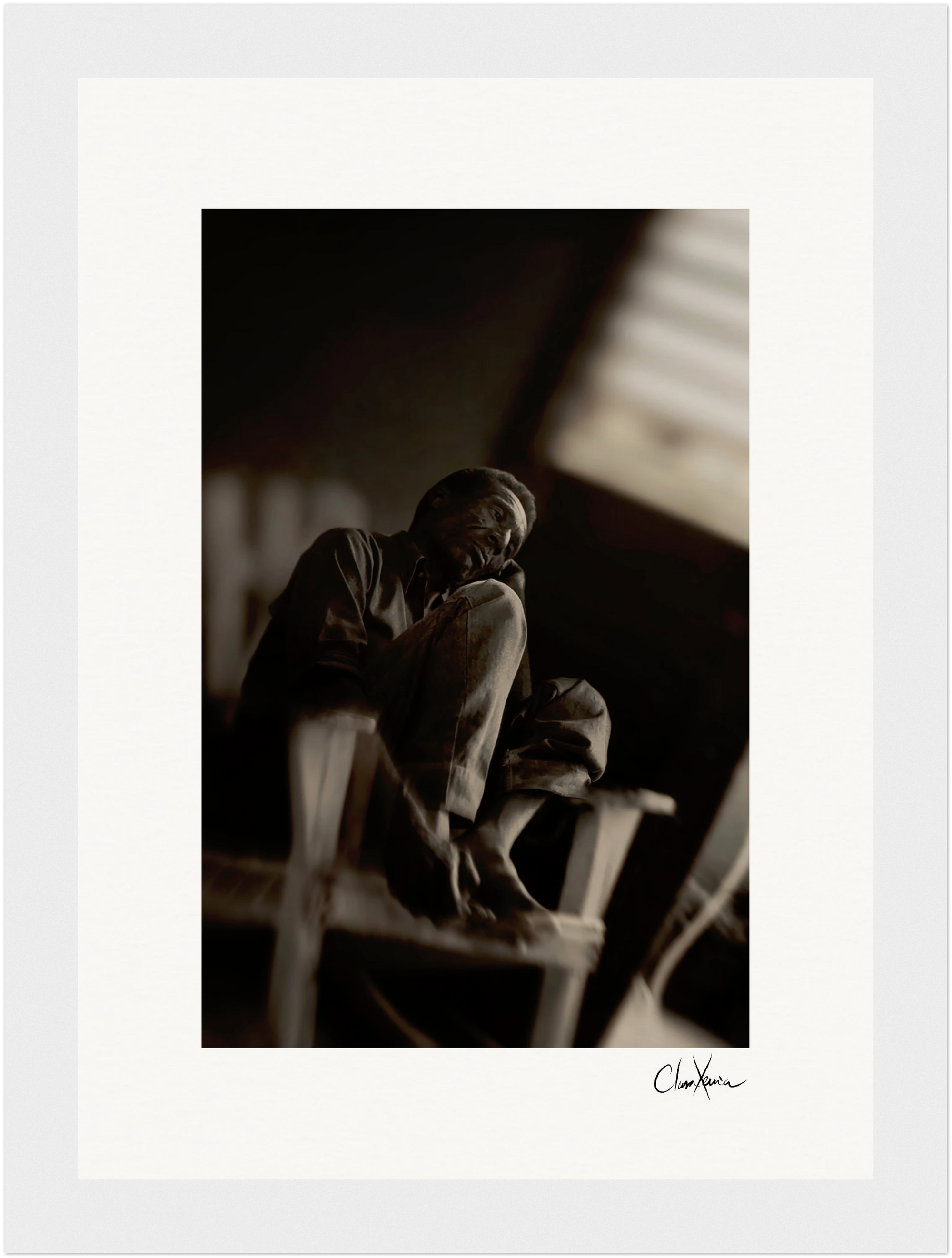 A black-and-white photo of a person sitting on a chair indoors, resting their chin on their hand with one leg pulled up. The blurred, dimly lit background enhances the conscious interior design, making it perfect as inspirational wall art.