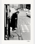 An elderly woman with a cane stands at the edge of a crosswalk on a cobblestone street, cars and pedestrians in the background. This black and white fine art print with a white border is perfect for mindful home decor.