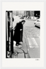 An elderly woman with a cane stands at the edge of a crosswalk on a cobblestone street, cars and pedestrians in the background. This black and white fine art print with a white border is perfect for mindful home decor.