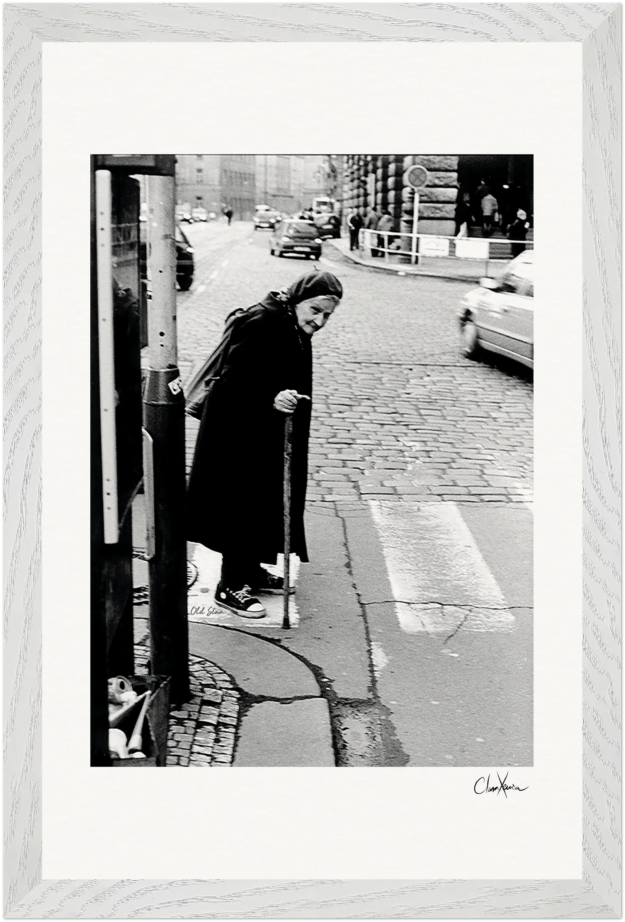 An elderly woman with a cane pauses at a cobblestone crosswalk, city cars passing by. Wearing a long coat and scarf, she is captured in black and white. This signed fine art print adds mindful home decor to any space.