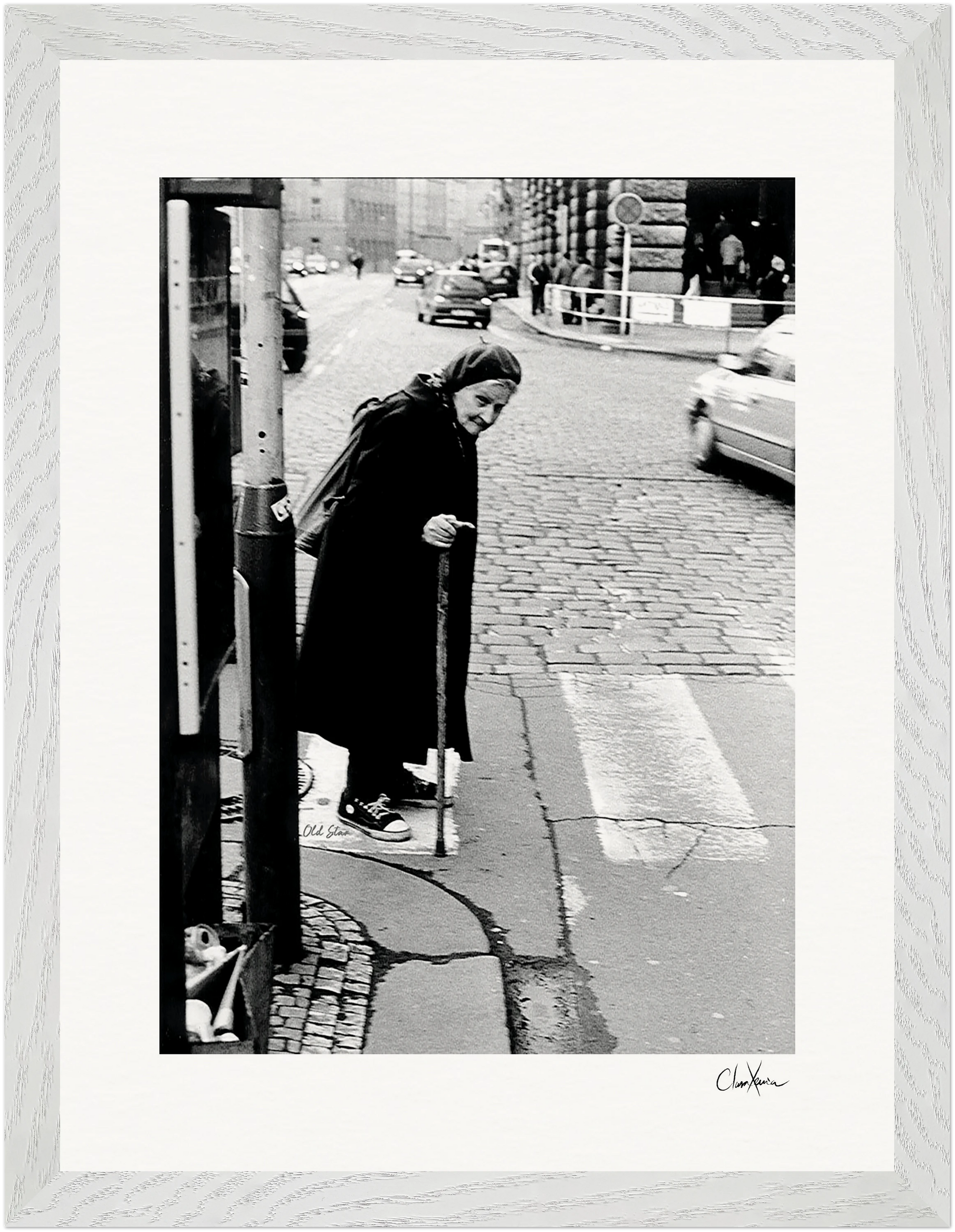 An elderly woman with a cane stands at a crosswalk on a cobblestone city street, wearing a long dark coat and headscarf. Cars appear in the background of this black and white fine art print, ideal as inspirational wall art.