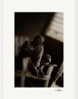 A man sits barefoot on a wooden chair, knees drawn to his chest, looking contemplative. This black and white fine art print has a subtle blur for a moody atmosphere—perfect as mindful home decor. A signature appears at the bottom.