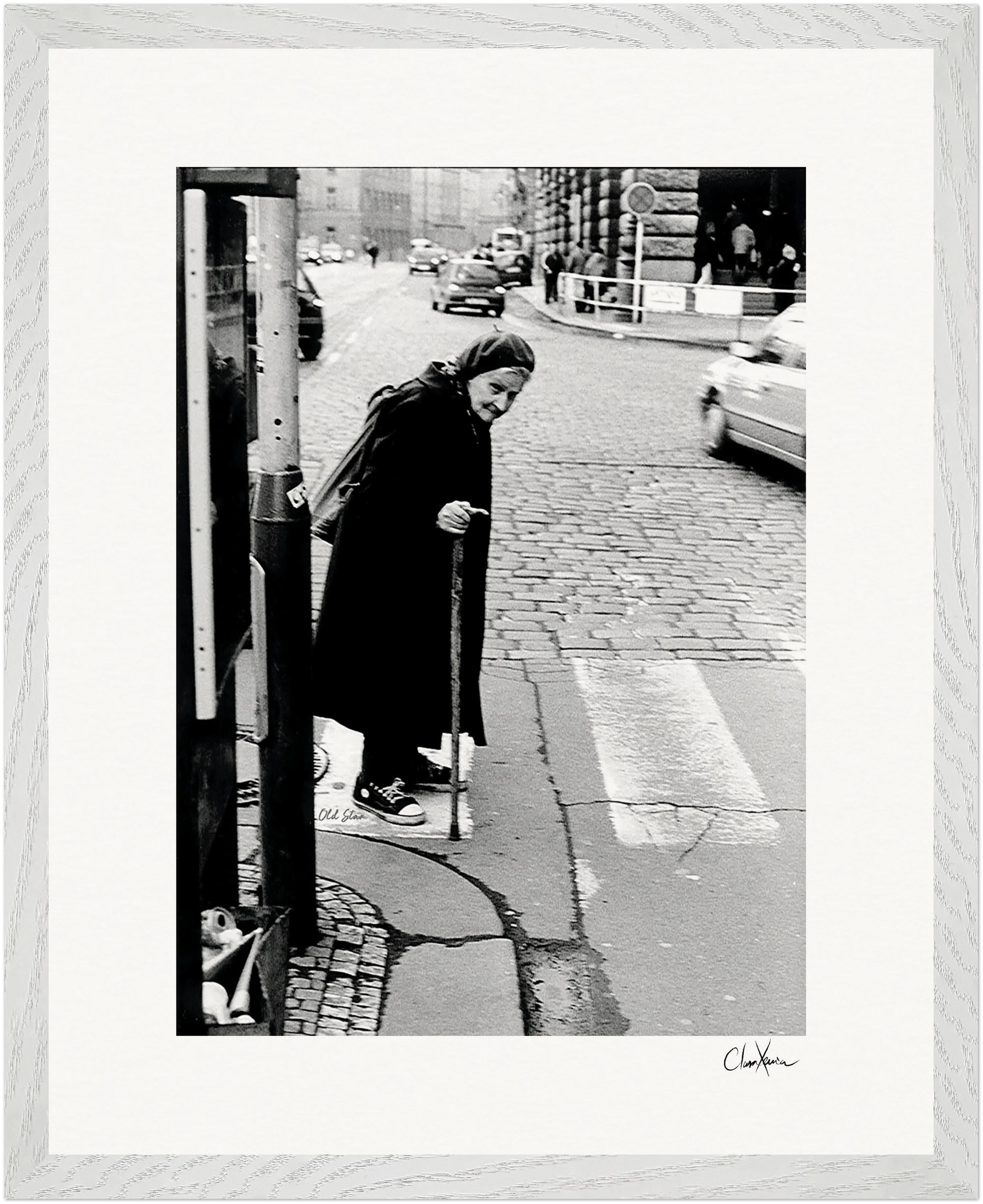 An elderly woman with a cane stands at the edge of a cobblestone street, ready to cross. She wears a dark coat and headscarf. Cars and buildings line the background. This black-and-white fine art print is presented in a gallery-quality frame.