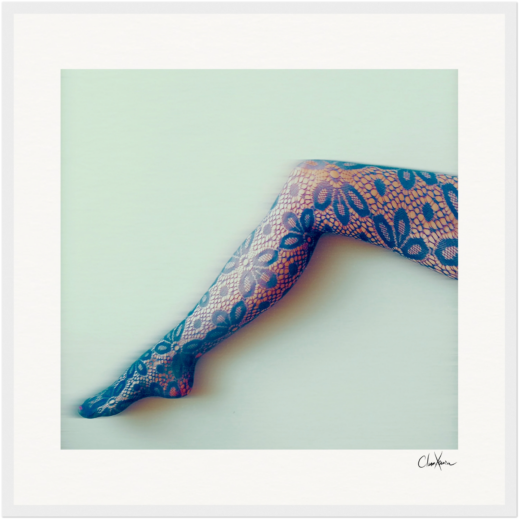A person's leg extended against a pale background, wearing black lace tights with an intricate floral and geometric pattern. This fine art print features a soft, high-contrast effect and a white border with a signature in the lower right corner.
