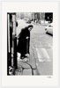 An elderly woman with a cane crosses a cobblestone city street at a crosswalk, wearing a dark coat and headscarf. This fine art print adds character and thoughtful beauty to any space, perfect for mindful home decor.