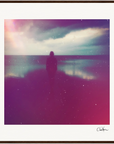 A silhouette of a person stands on a beach facing the water under a cloudy sky, with a soft pink and purple filter. Small white specks are scattered across the photo, making it perfect as framed wall art for mindful home decor.