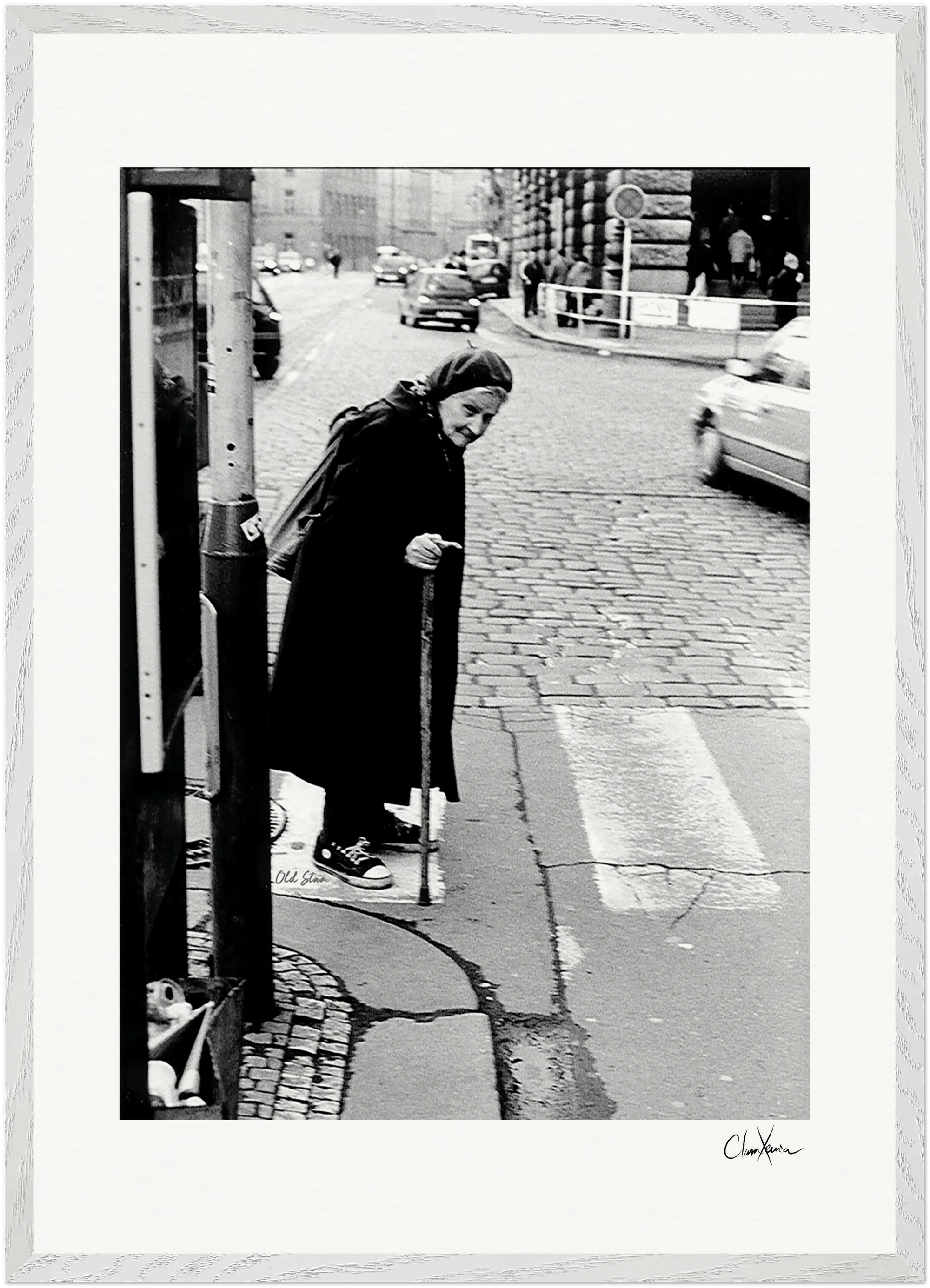 An elderly woman in a headscarf and dark coat uses a cane to cross a city street, with cars in the background. This black and white image makes striking framed wall art, perfect for mindful home decor or gallery-quality framing.
