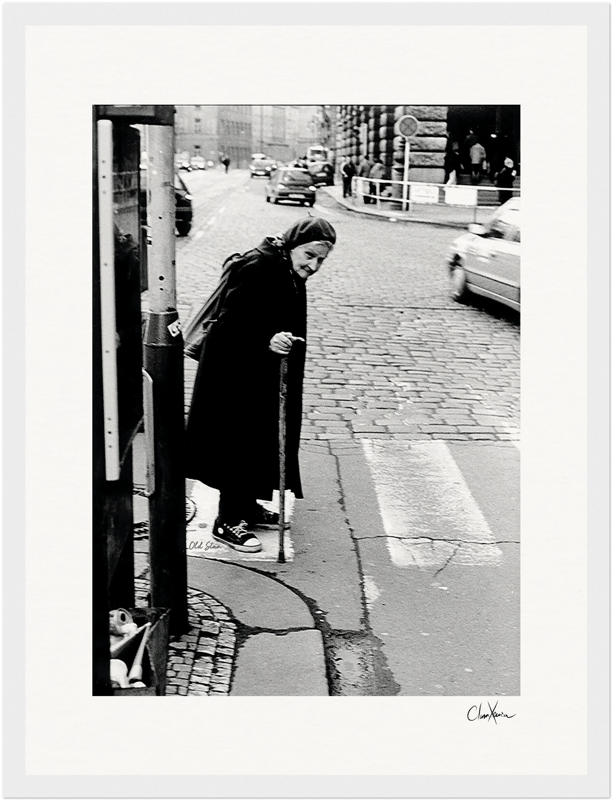 An elderly woman with a cane stands at the edge of a crosswalk on a cobblestone city street, wearing a long dark coat and scarf; cars and buildings are visible in the background in this framed wall art.