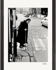 An elderly woman using a cane crosses a cobblestone street at a crosswalk in an urban setting, captured in black and white and displayed in a gallery-quality frame—an inspiring choice for mindful home decor.