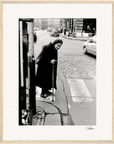 An elderly woman in a long coat and headscarf uses a cane while stepping onto a cobblestone street at a crosswalk; cars and buildings line the background. This black and white photo makes thoughtful inspirational wall art for mindful home decor.