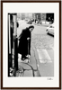 An older woman with a cane crosses a cobblestone street at a crosswalk in this black and white photo. Wearing a dark coat and headscarf, she moves past cars and pedestrians—perfect as framed wall art for conscious interior design.