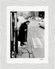 An elderly woman with a cane stands at a crosswalk on a cobblestone street, wearing a long dark coat. Cars and buildings are in the background. This black-and-white photo in a white wood frame adds mindful home decor as striking framed wall art.