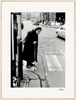 An elderly woman in a headscarf and long coat uses a cane to cross a cobblestone city street near a telephone booth. The black and white image makes striking framed wall art for conscious interior design. Cars are visible in the background.