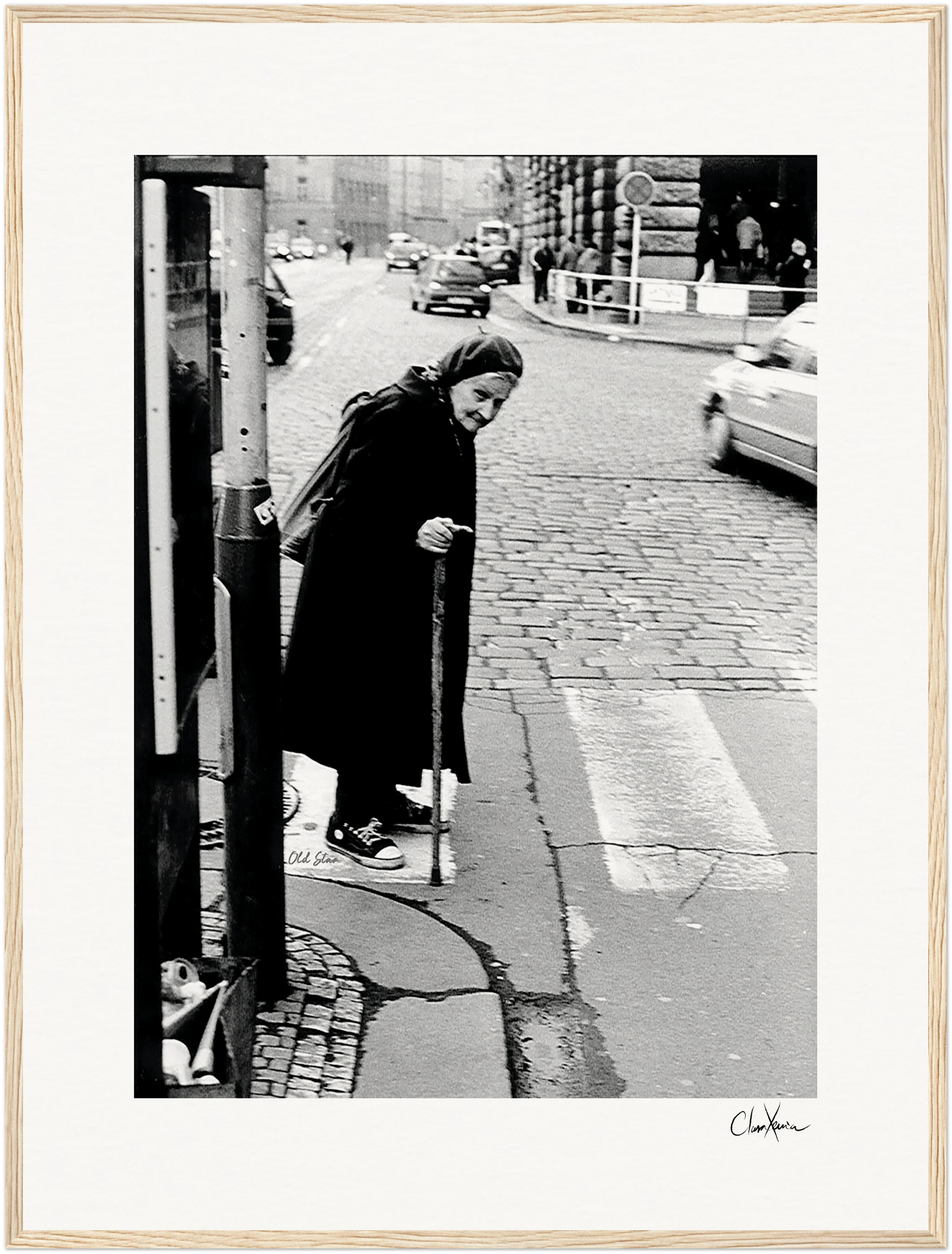 An elderly woman in a headscarf and long coat uses a cane to cross a cobblestone city street near a telephone booth. The black and white image makes striking framed wall art for conscious interior design. Cars are visible in the background.