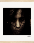 A grainy, close-up image of a person's face with dark hair partially covering it, set against a plain light background and framed with a light wooden frame. Inspirational wall art, this fine art print features a signature in the lower right corner of the border.