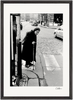 An elderly woman with a cane crosses a cobblestone street at a crosswalk, dressed in a long dark coat and headscarf. Cars and buildings are visible in the black-and-white image, ideal as a fine art print for mindful home decor.