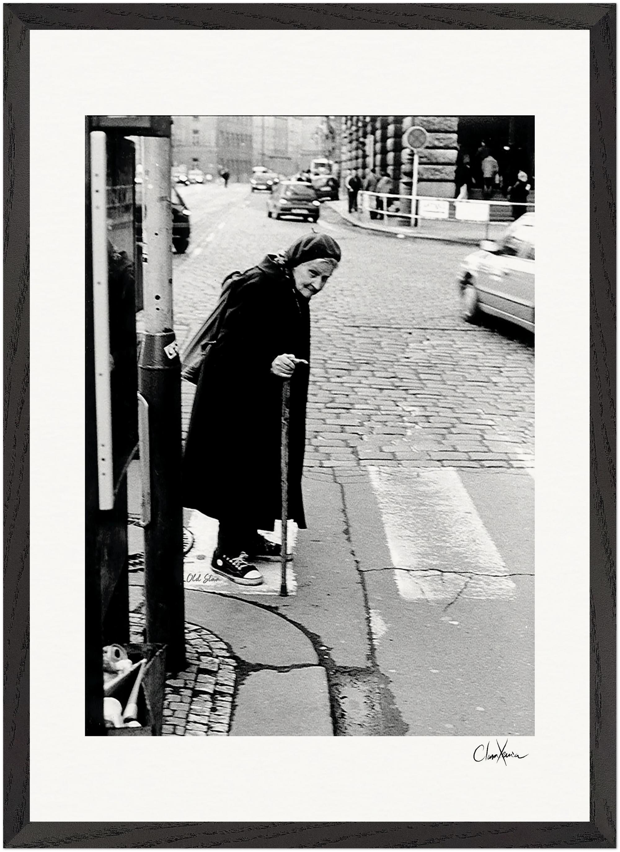 An elderly woman with a cane crosses a cobblestone street at a crosswalk, dressed in a long dark coat and headscarf. Cars and buildings are visible in the black-and-white image, ideal as a fine art print for mindful home decor.