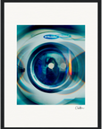 A framed fine art print of a washing machine door with light reflections and blue tones, featuring the Electrolux brand and an abstract reflection of a person. This inspirational wall art is signed in the bottom right corner.