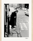 An elderly woman wearing a headscarf and long coat walks with a cane on a cobblestone city street, pausing at a crosswalk. Her mindful presence evokes the quiet inspiration often found in conscious interior design and inspirational wall art.