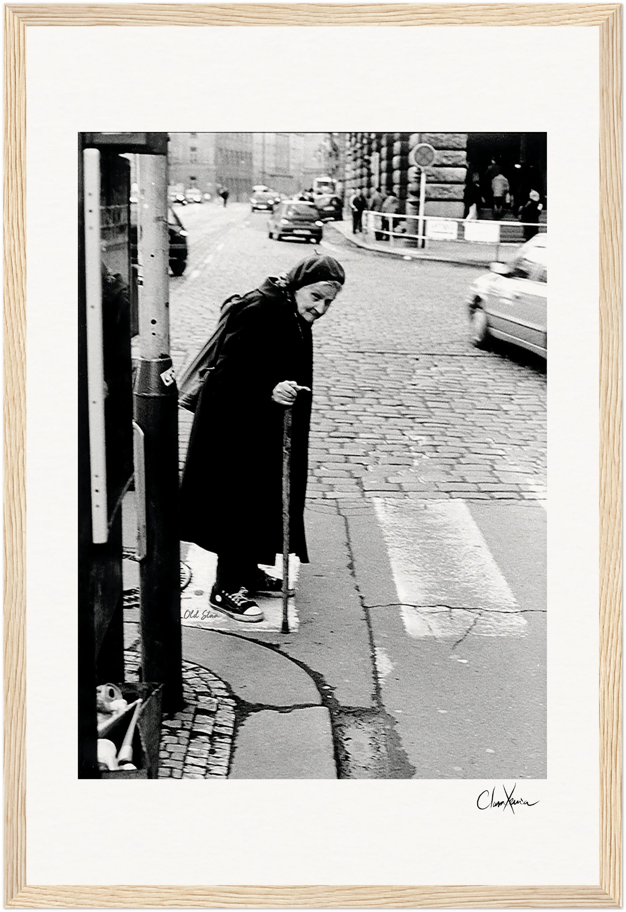 An elderly woman wearing a headscarf and long coat walks with a cane on a cobblestone city street, pausing at a crosswalk. Her mindful presence evokes the quiet inspiration often found in conscious interior design and inspirational wall art.