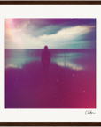 A silhouette of a person stands facing calm water under a cloudy sky, with pink and purple light effects giving the photo a dreamy look. This inspirational wall art is framed and signed in the bottom right corner—perfect for mindful home decor.
