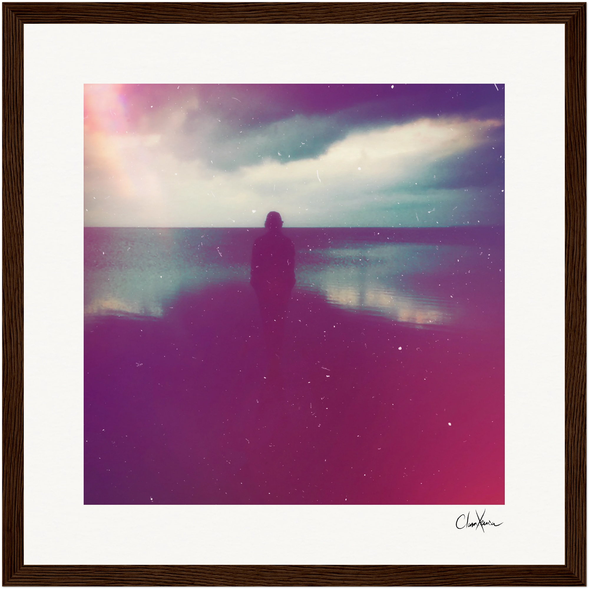 A silhouette of a person stands facing calm water under a cloudy sky, with pink and purple light effects giving the photo a dreamy look. This inspirational wall art is framed and signed in the bottom right corner—perfect for mindful home decor.