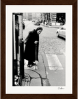 An elderly woman in a dark coat and headscarf uses a cane while crossing a cobblestone street at a crosswalk. Cars and buildings frame the black and white scene, making it perfect as inspirational wall art for conscious interior design.
