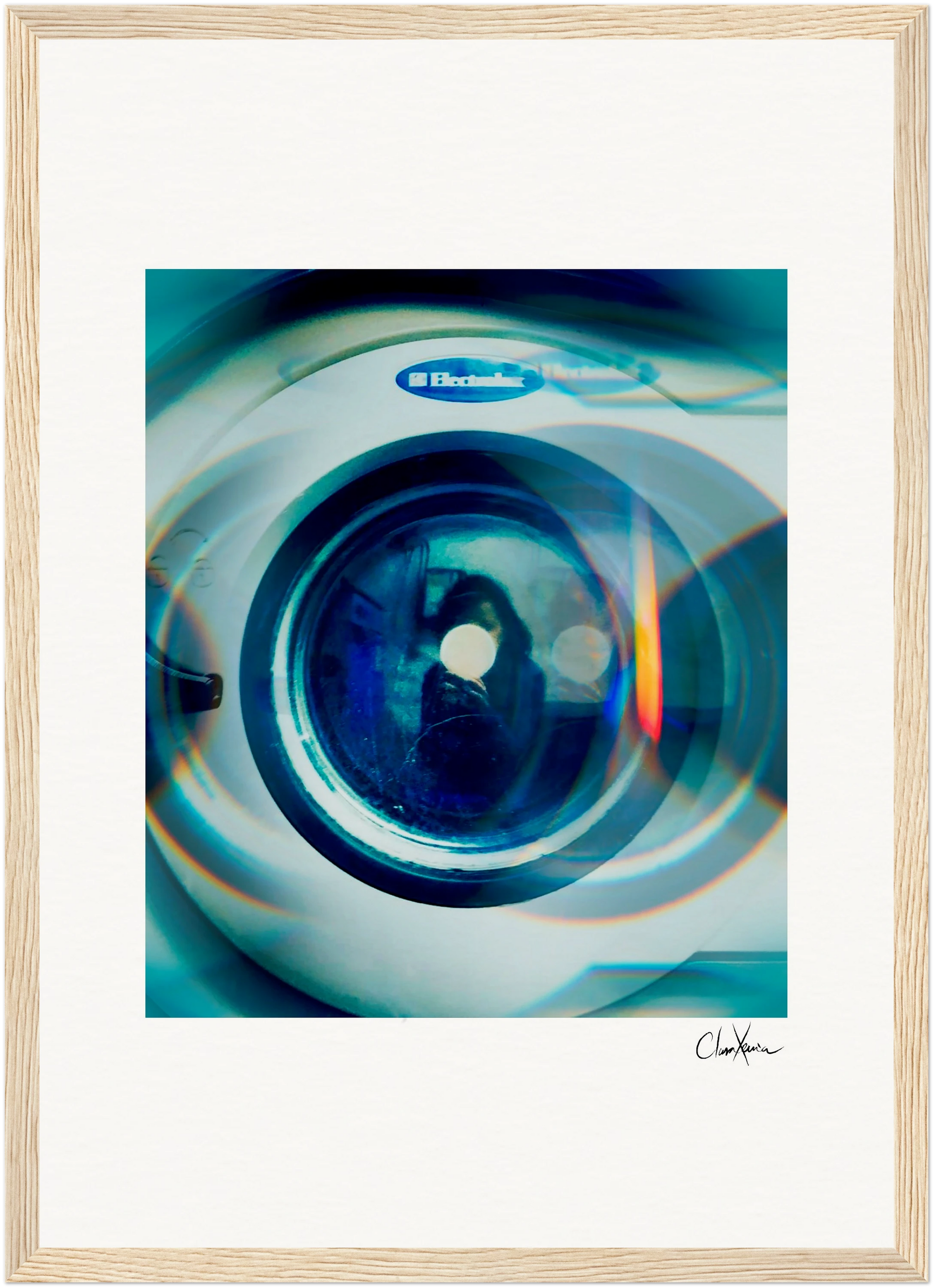 A close-up view of the circular door of an Electrolux washing machine with blue and orange light reflections, elegantly presented as framed wall art. The image is set in a gallery-quality frame with a signature at the bottom right.