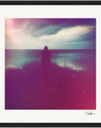 A silhouetted person stands on a beach facing the water under a cloudy sky, with pink and purple filters giving the image a dreamy, ethereal effect. Perfect as inspirational wall art for conscious interior design. A signature appears in the bottom right corner.