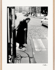 An elderly woman in a headscarf and dark coat uses a cane while crossing a cobblestone street at a crosswalk, surrounded by cars and buildings—an evocative scene perfect for inspirational wall art or mindful home decor.