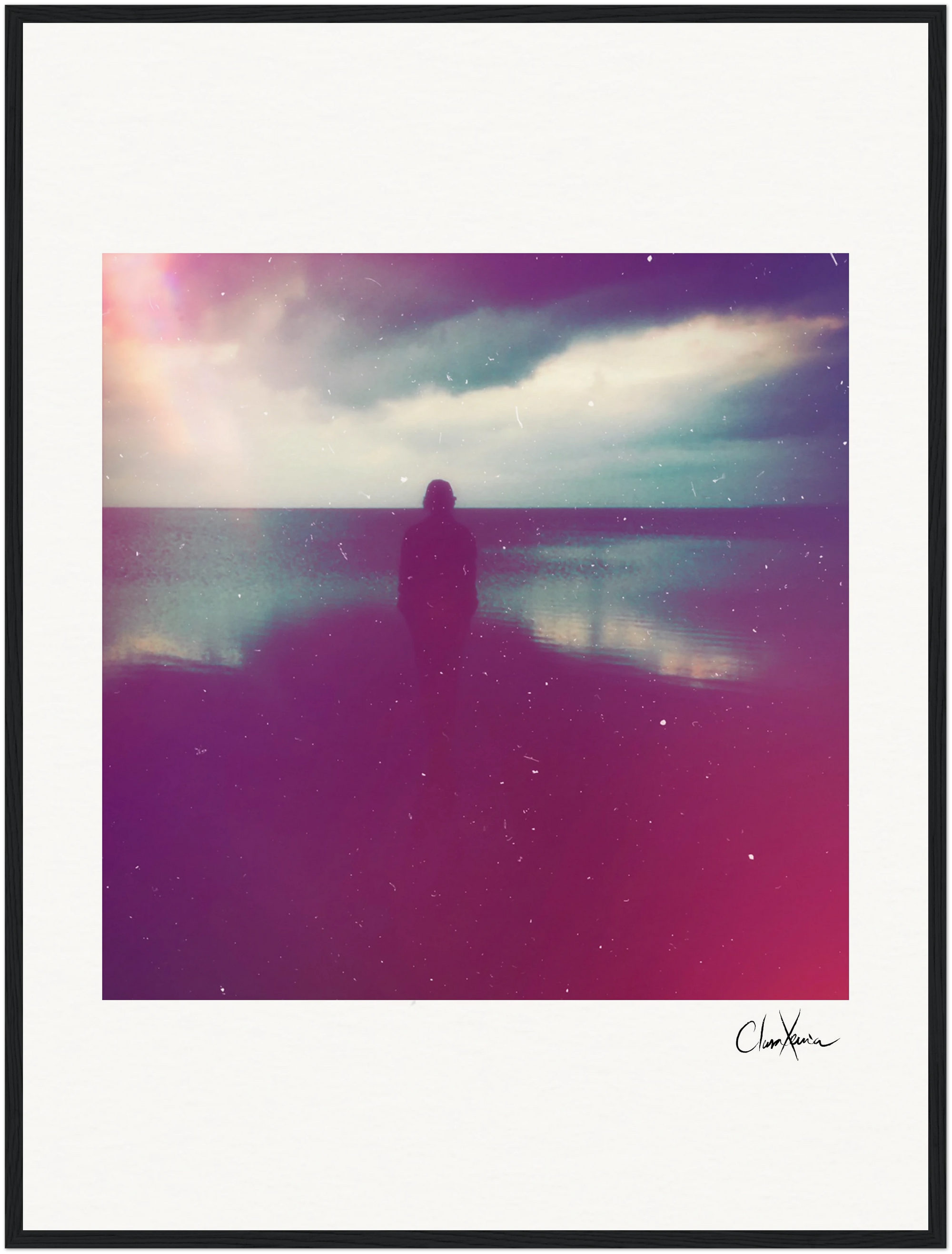 A silhouetted person stands by a calm body of water under a cloudy sky, with soft pink and purple hues creating a dreamy atmosphere. This fine art print features a grainy texture and white border, perfect for conscious interior design or inspirational wall art.