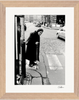 An elderly woman with a cane stands at a crosswalk on a cobblestone street. Cars and buildings form the background. This black and white fine art print is beautifully framed with light brown wood, perfect for mindful home decor.