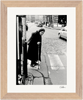 An elderly woman with a cane stands at a crosswalk on a cobblestone street. Cars and buildings form the background. This black and white fine art print is beautifully framed with light brown wood, perfect for mindful home decor.
