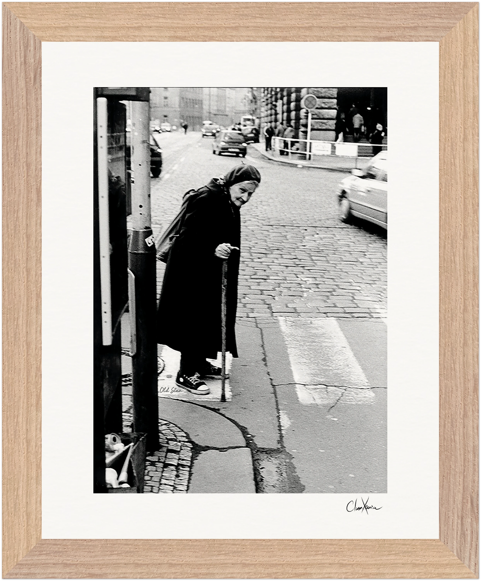 An elderly woman with a cane stands at a crosswalk on a cobblestone street. Cars and buildings form the background. This black and white fine art print is beautifully framed with light brown wood, perfect for mindful home decor.