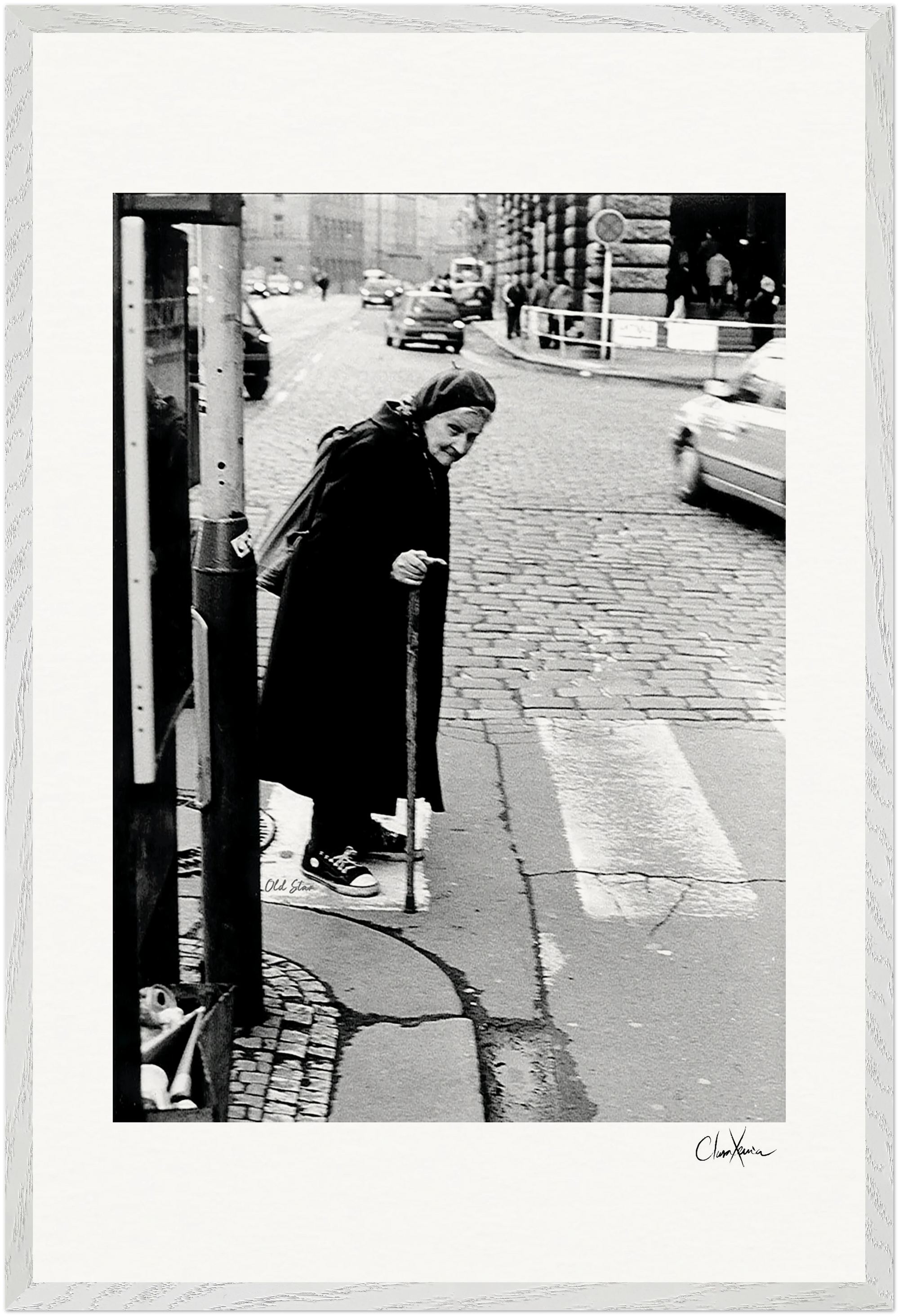 An elderly woman with a cane walks slowly across a cobblestone street at a crosswalk. Cars are visible in the background; she wears a long coat and headscarf. This black and white photo creates inspirational wall art perfect for mindful home decor.