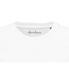 Close-up of the neckline of a plain white T-shirt, featuring "Abundance" in blue script on the inside collar. This mindful apparel piece includes a small brown size tag on the right, reflecting conscious fashion values.