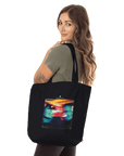 A woman with long brown hair holds a black tote bag made from organic material over her shoulder. The tote features a colorful, abstract design. She is wearing a short-sleeved olive green shirt and stands against a plain white background.