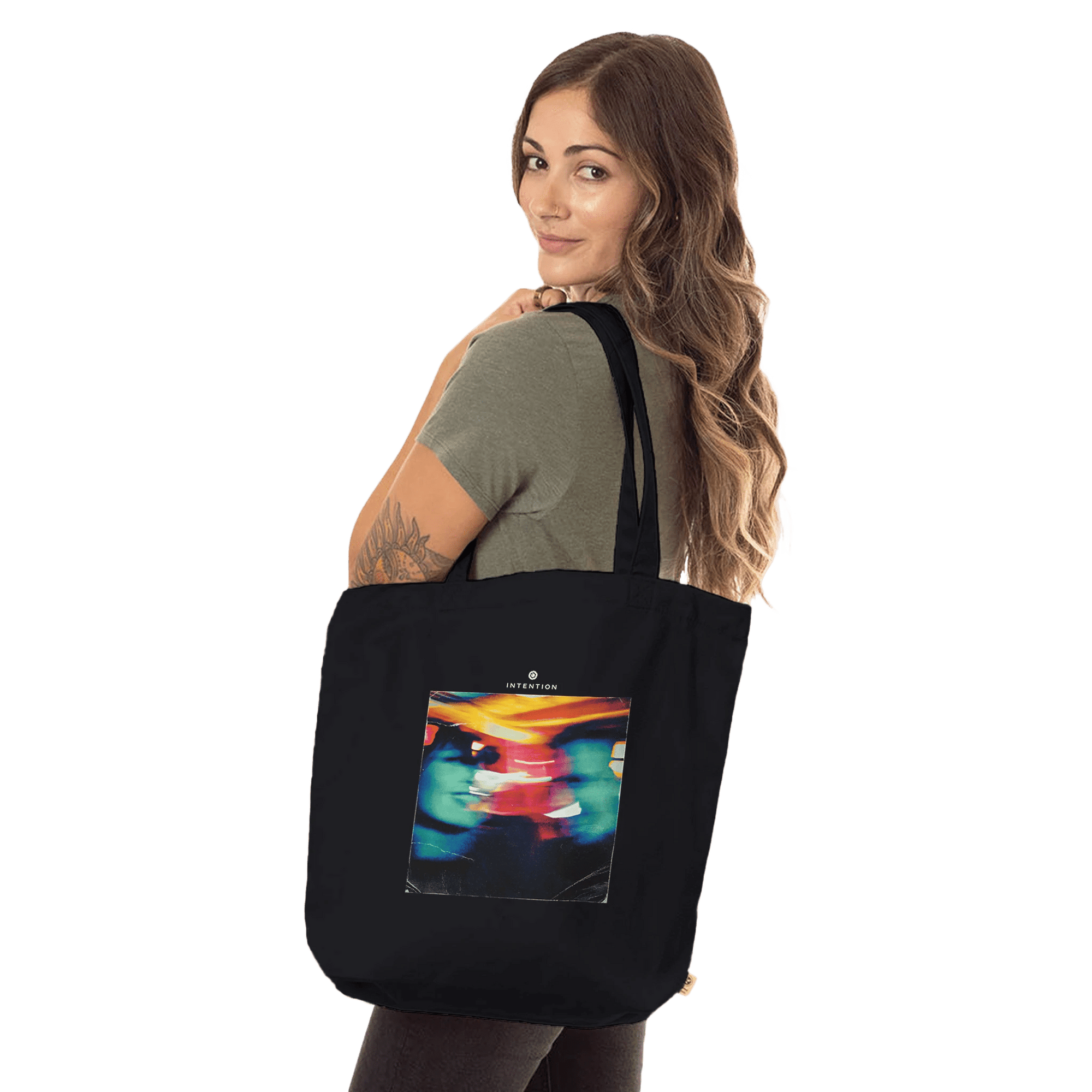 A woman with long brown hair holds a black tote bag made from organic material over her shoulder. The tote features a colorful, abstract design. She is wearing a short-sleeved olive green shirt and stands against a plain white background.